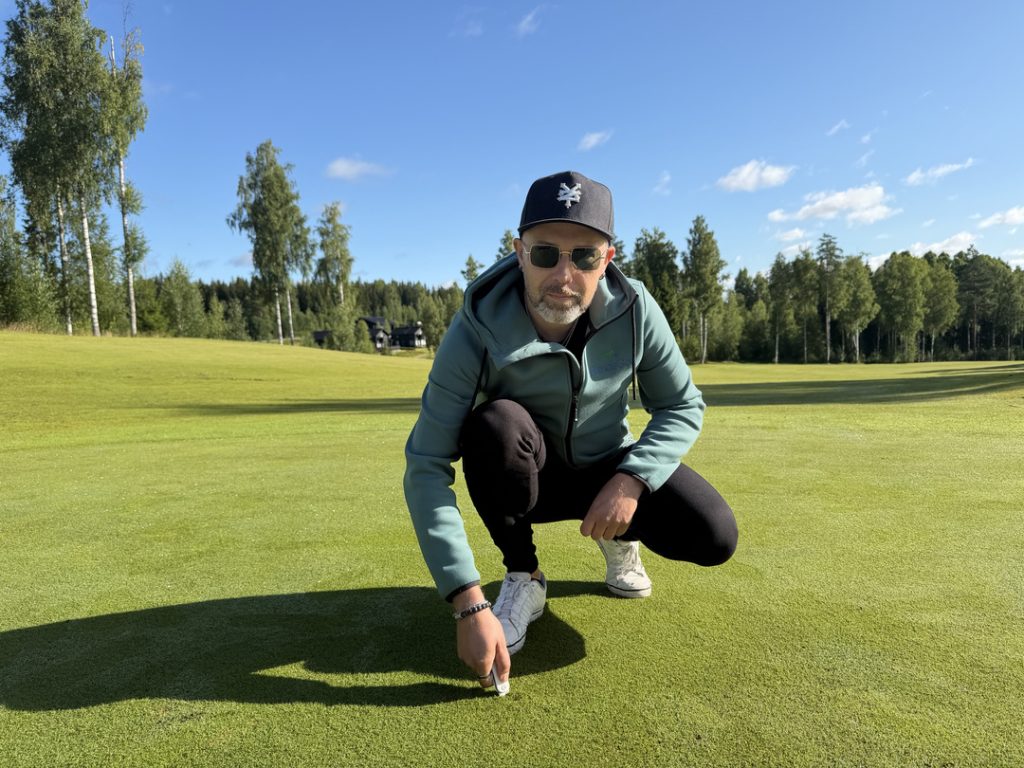 Himos Golfin vihreällä ja aurinkoisella golfkentällä henkilö painaa greenihaarukkaa viheriölle ja katsoo kameraan aurinkolasit päässään.
