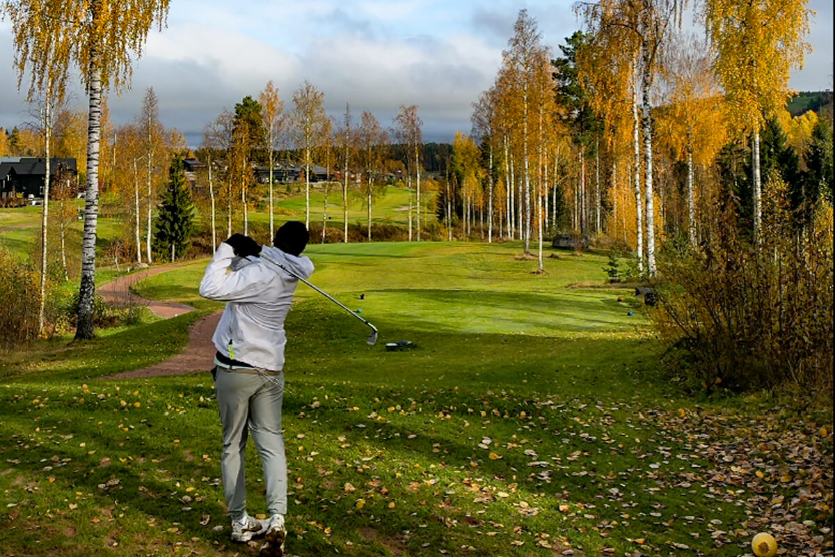 Valkoisessa takissa oleva golffari lyö syysmaisemassa palloa Himos Golfissa. Puolipilvinen sää, puut ja syksyn ruska on voimakas.