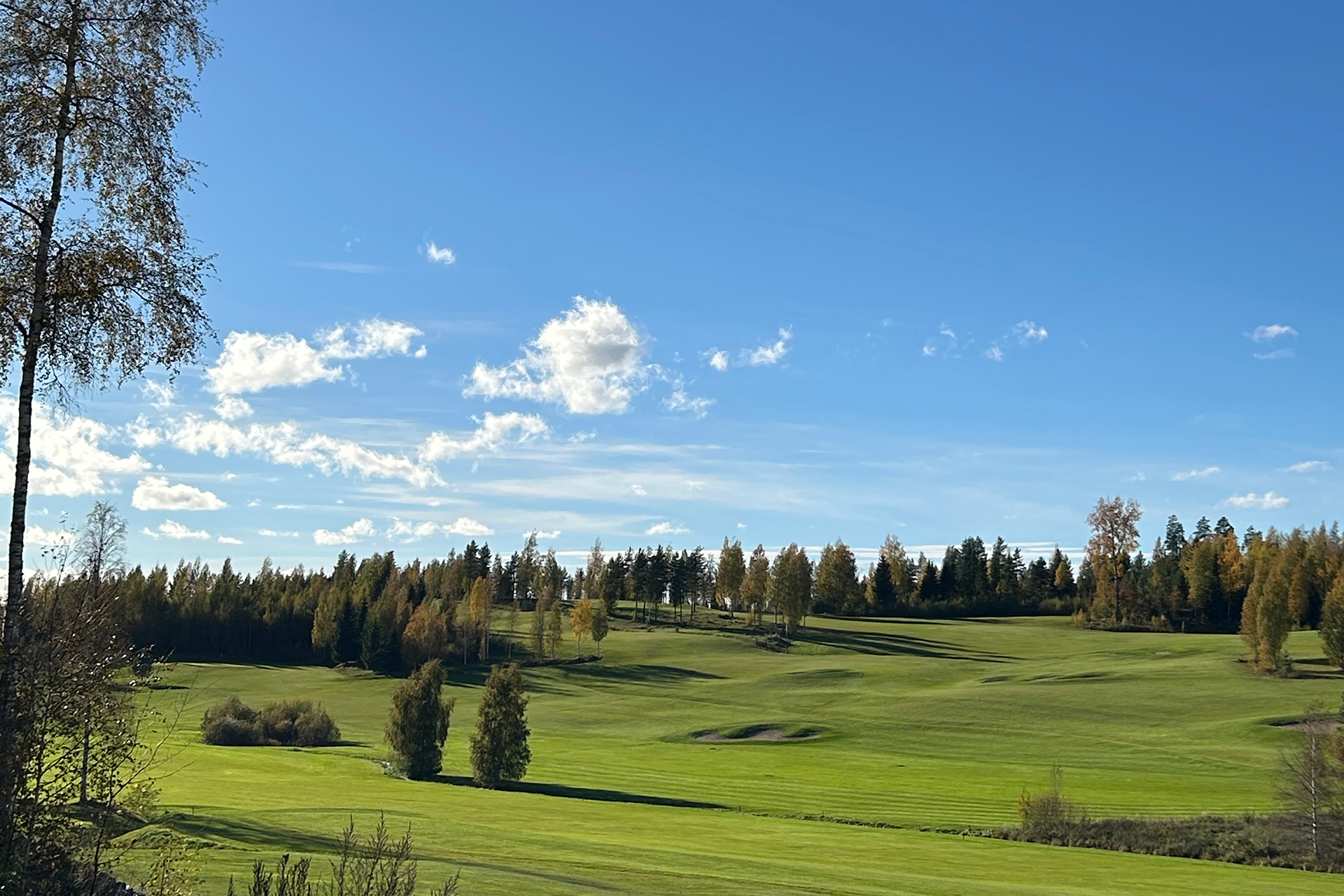 Syksyinen ja aurinkoinen maisemakuva Himos Golfista. Kenttä on vihreä, mutta taustalla puut ovat ruskan värittämät.